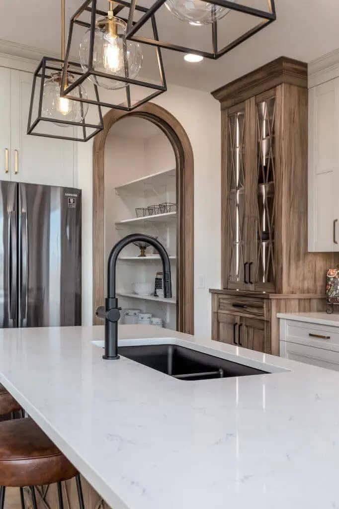 Modern kitchen with a white marble island, black faucet, and wooden cabinetry. Pendant lights hang above; stainless steel fridge in the background. Cozy and elegant.