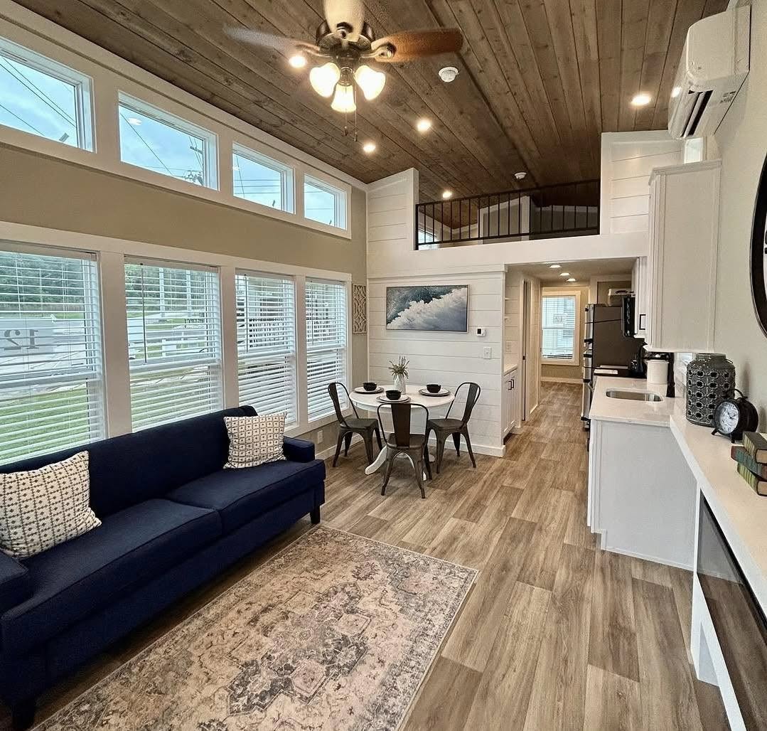 Modern living space with a vaulted wooden ceiling, large windows, a blue couch, a round dining table, and a compact kitchen. Bright and cozy.