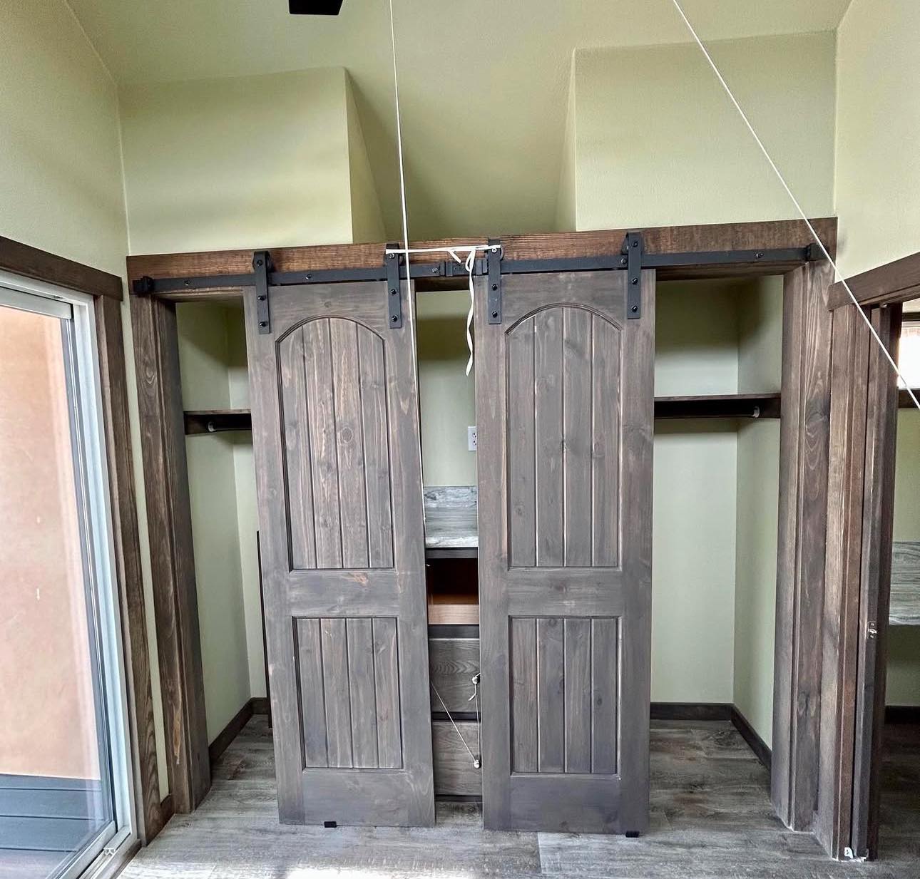 Wooden sliding barn doors with arched panels cover a closet, set in a room with light green walls and wood vinyl flooring, conveying rustic charm.
