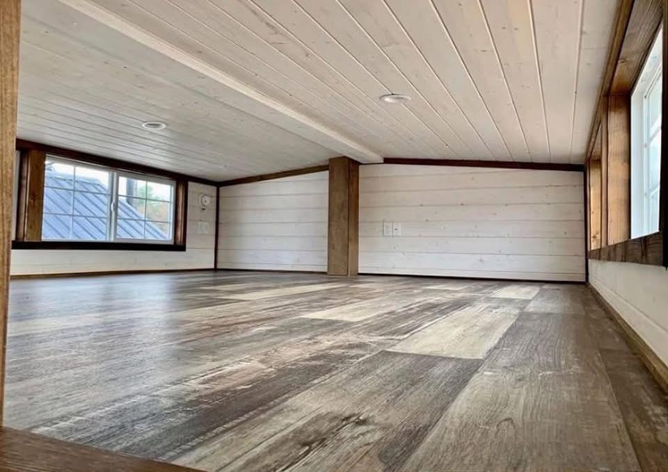 Spacious room with white wooden walls and ceiling, featuring dark wood accents and a large window, creating a bright and airy atmosphere.
