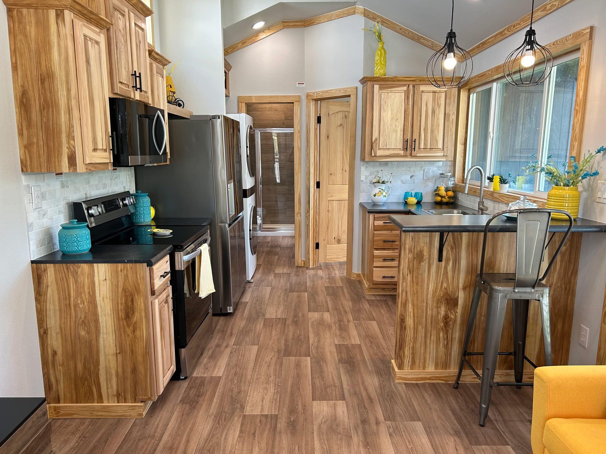 A modern kitchen with natural wood cabinets, black countertops, and stainless steel appliances. Bright accents include yellow decor and vibrant plants.