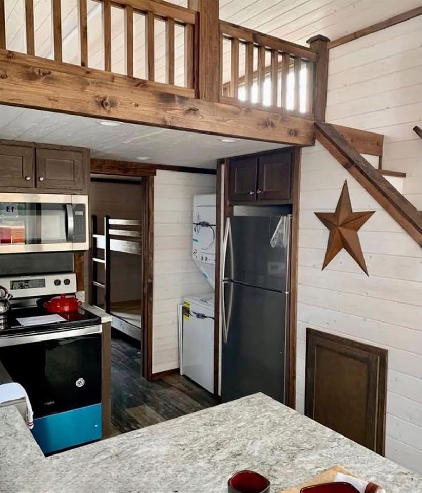 Cozy kitchen scene with wood finishes, a star on the wall, a loft railing above, bunk beds, and a compact washer-dryer next to the fridge.