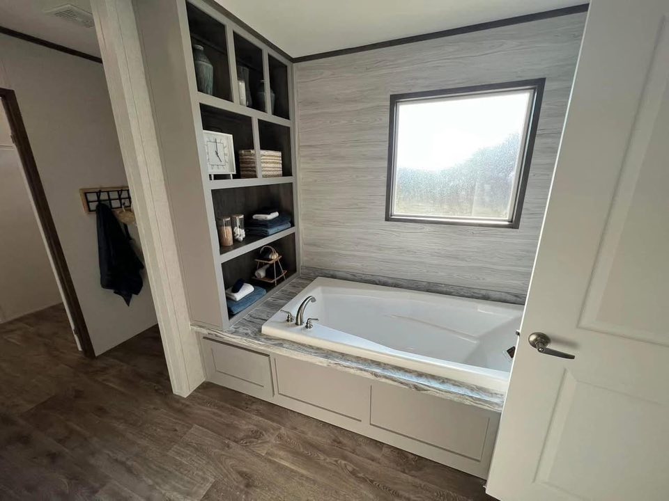 A modern bathroom with a sunken bathtub, gray wood paneling, open shelves with decor, and a foggy window. The room feels serene and organized.