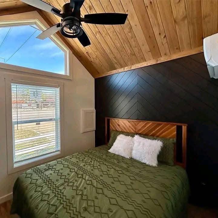 Cozy bedroom with wood-paneled ceiling, black accent wall, and green bedspread. Sunlight streams through a large window, illuminating the space.