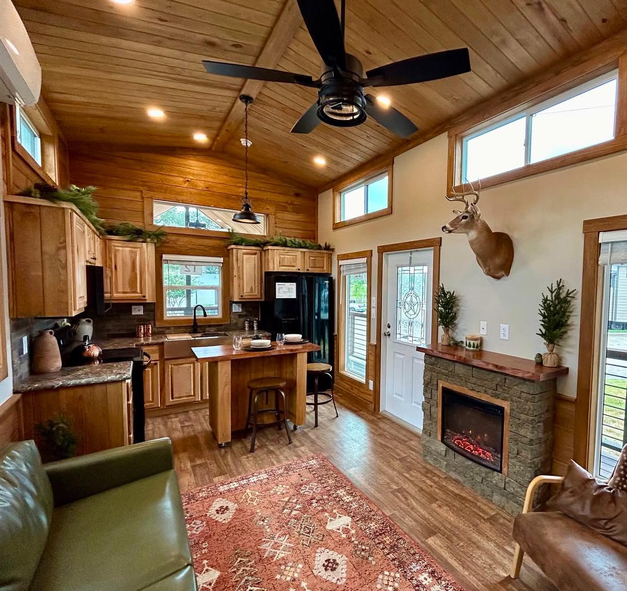 Cozy rustic cabin interior with wooden ceiling, kitchen, and small living area. Features include a deer mount, electric fireplace, and a red patterned rug. Warm and inviting atmosphere.