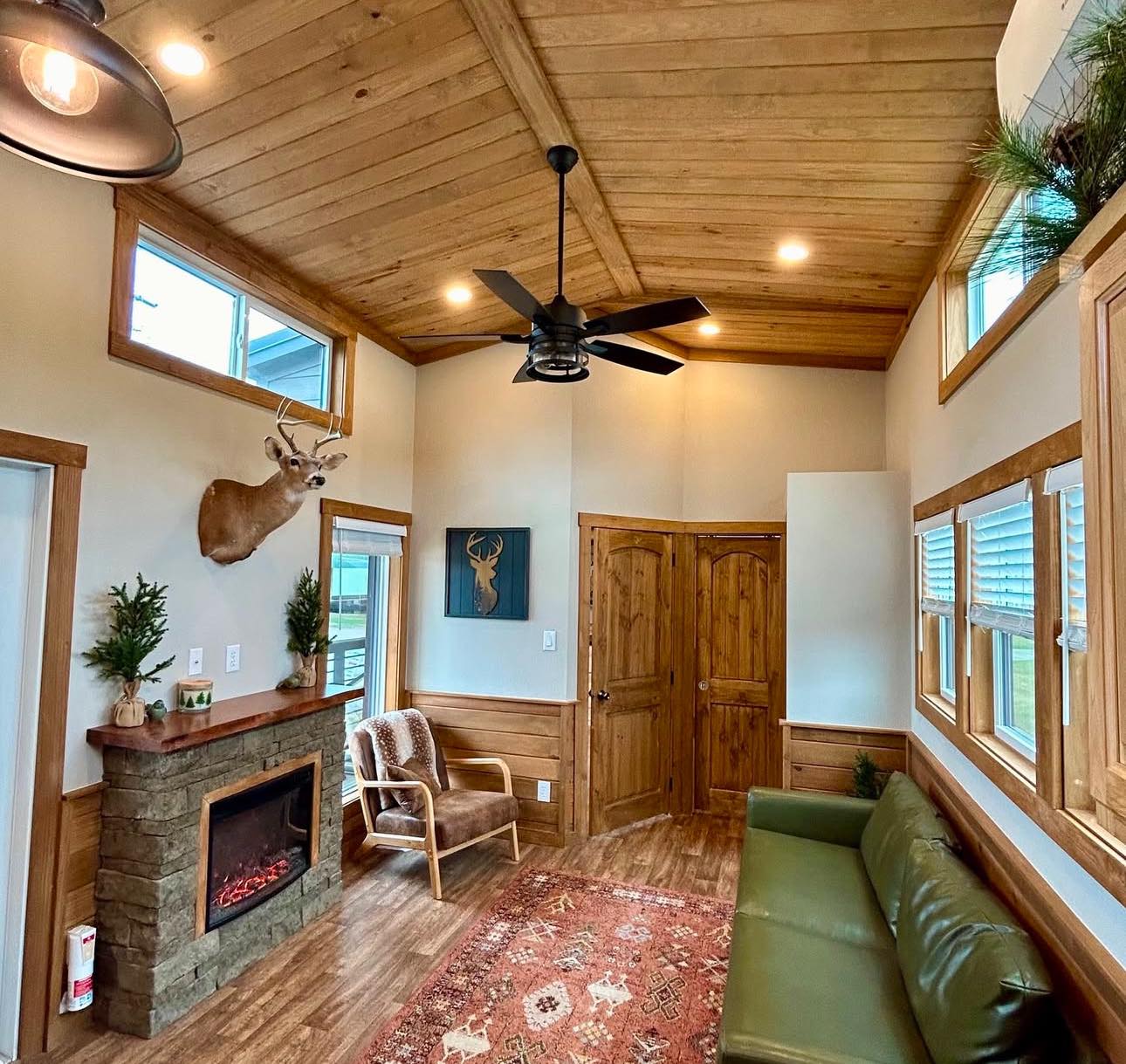 Cozy cabin room with wooden ceiling, fireplace, and armchair. A deer head mount and rustic decor add to the warm, inviting atmosphere.
