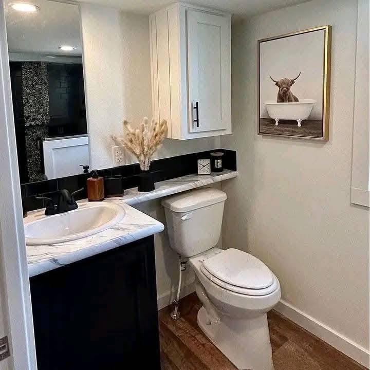 A small bathroom with a toilet, a black-cabinet sink, and decorative elements. A quirky cow-in-bathtub artwork adds humor above the toilet.