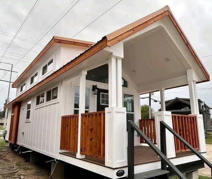 Charming tiny house with a gabled roof, white siding, and a wooden porch. Large windows enhance the cozy, welcoming vibe under a cloudy sky.