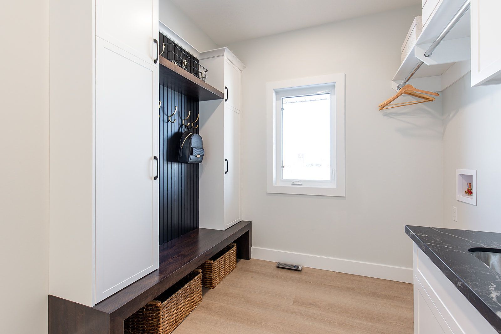 A tidy mudroom with white cabinets, wooden bench, and wicker baskets underneath. A black backpack hangs on hooks. Bright window, calm atmosphere.