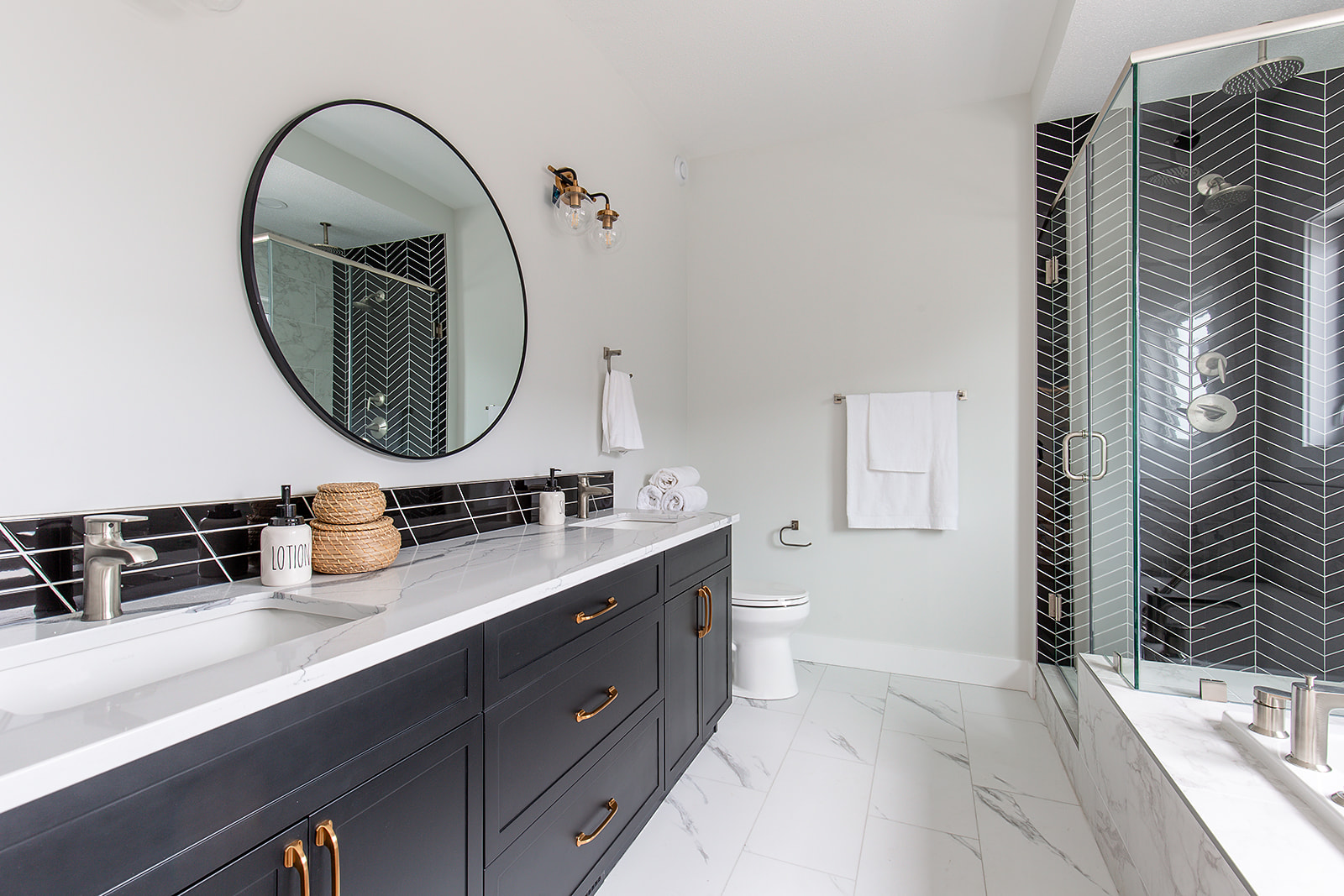 Modern bathroom with sleek black vanity, round mirror, and glass shower. White tiles and black herringbone pattern add contrast and elegance.