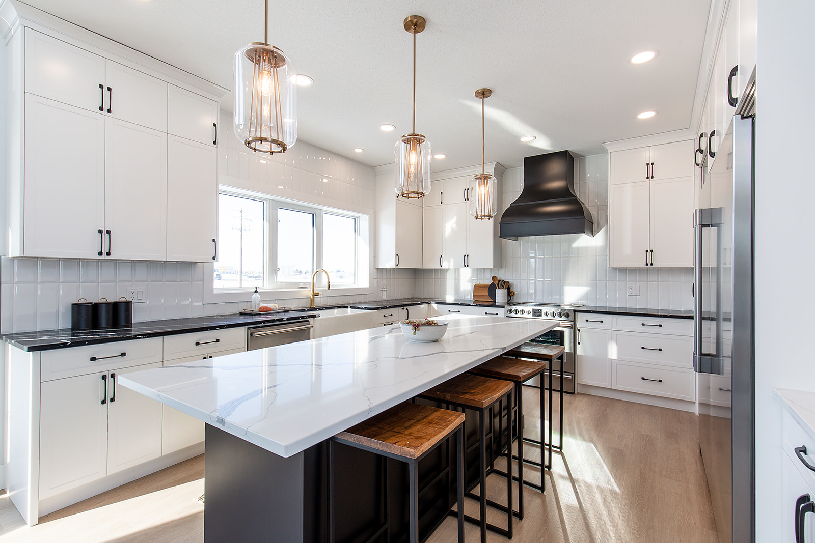 Modern kitchen with white cabinets, large island with wood stools, and pendant lights. Black range hood adds contrast, creating a sleek, inviting space.