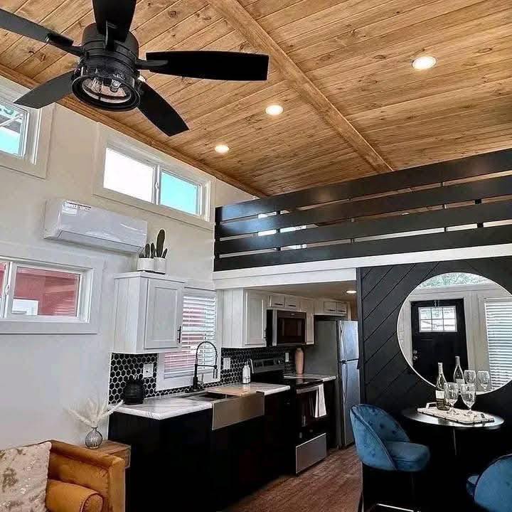 Modern tiny home interior with wood ceiling and black fan. Features include a loft, sleek kitchen, round mirror, and cozy dining area with blue chairs.