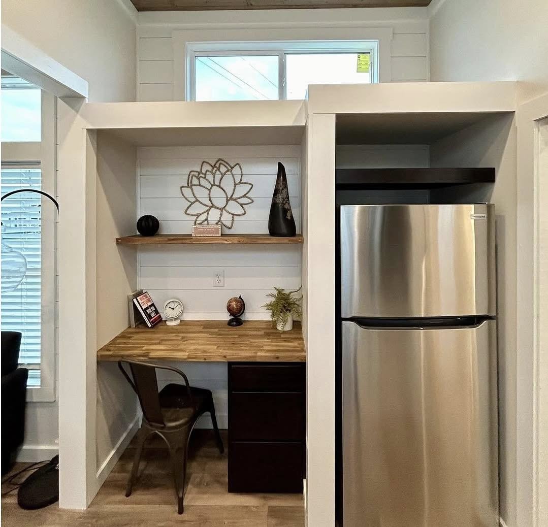 A cozy nook with a wooden desk, chair, and shelf adorned with decor. A stainless steel fridge stands to the right. The space feels modern and inviting.
