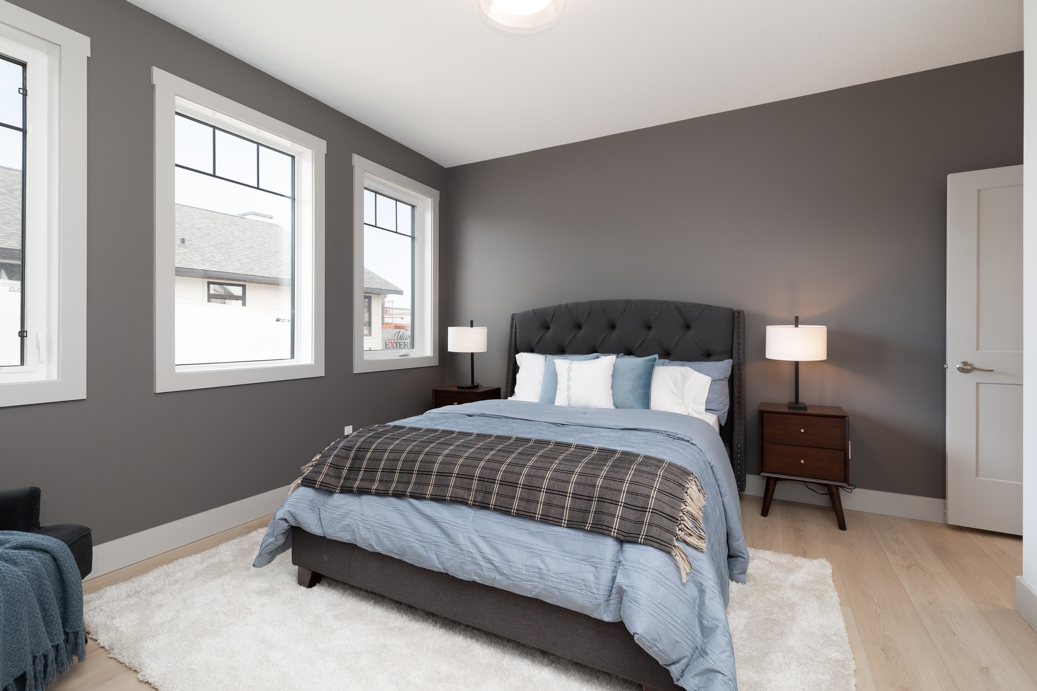 A cozy bedroom with gray walls features a plush bed with blue and white bedding and a plaid throw. Three large windows, two nightstands with lamps, and a soft white rug complete the serene atmosphere.