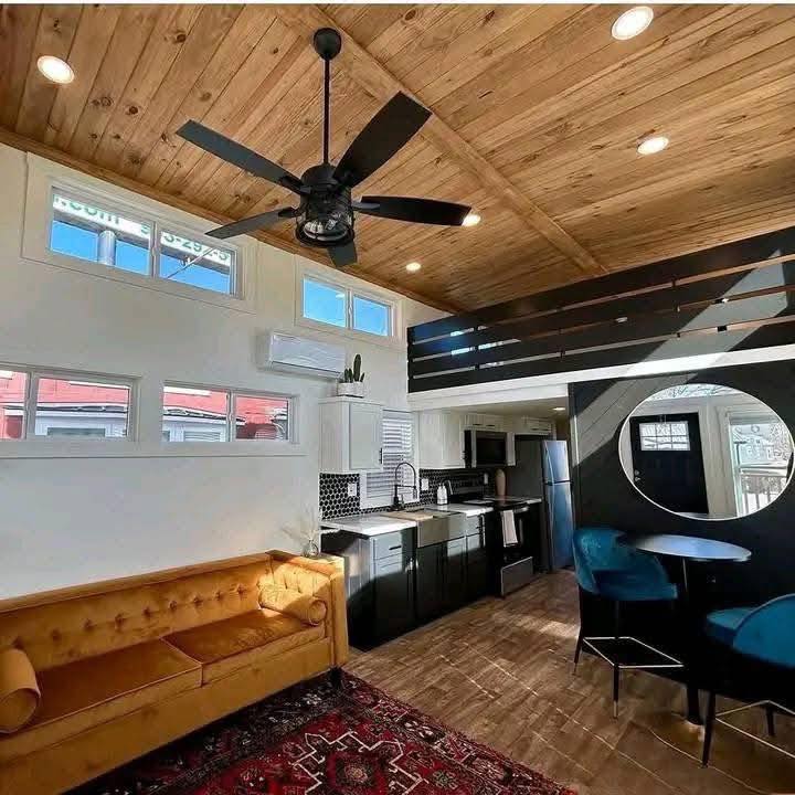 Stylish living space with a wooden ceiling and fan, featuring a mustard sofa, red carpet, modern kitchen, and circular wall art with blue chairs.