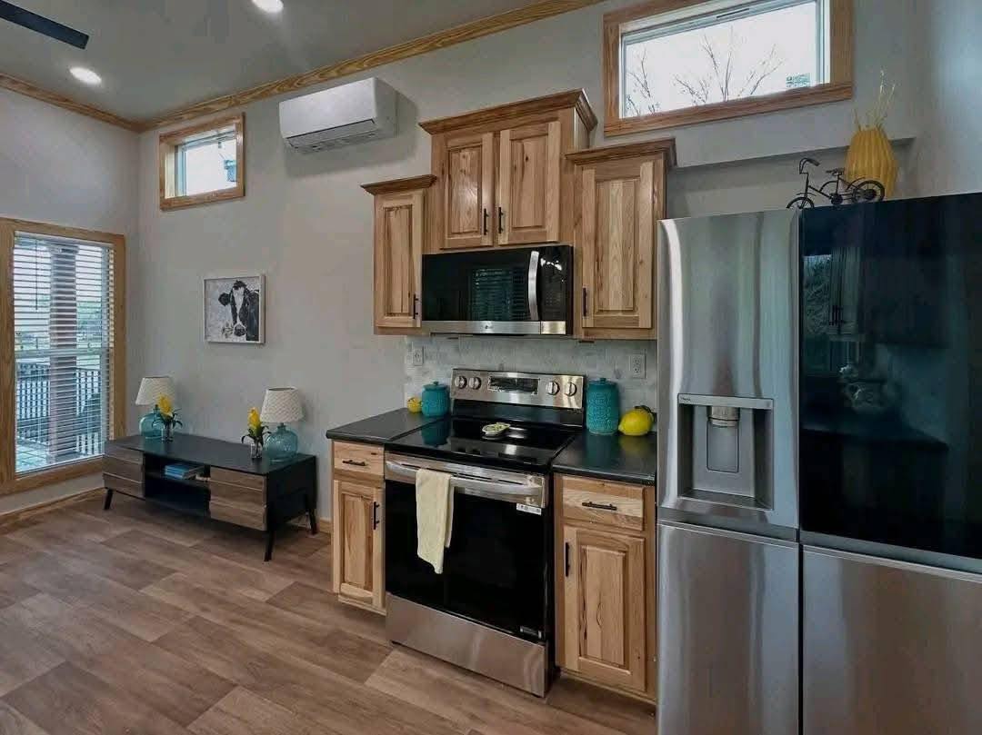 Bright kitchen with wooden cabinets, microwave, and stainless steel appliances. Light wood flooring, large windows, and modern decor create a cozy ambiance.