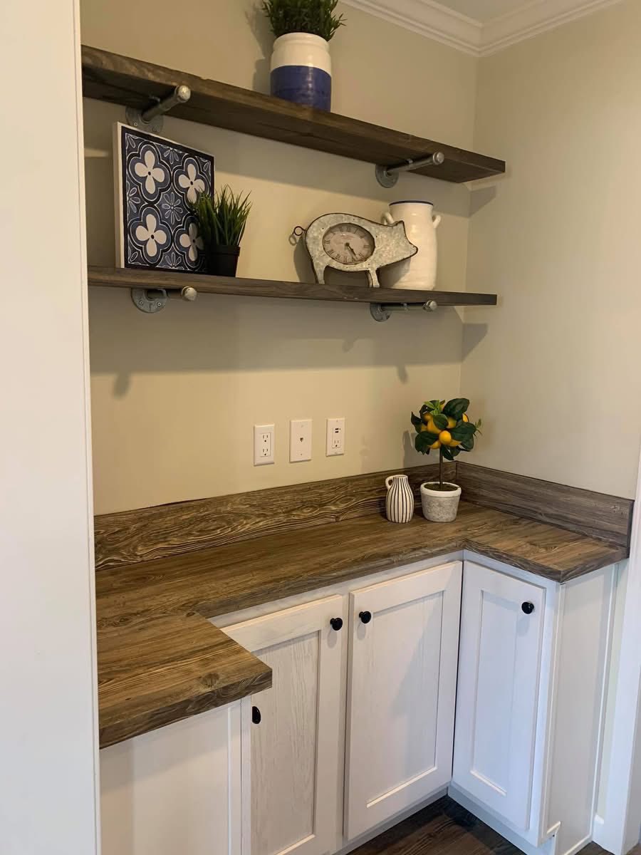 Small kitchen nook with two wooden shelves displaying a blue vase, plants, a decorative plate, and a pig-shaped clock. A rustic countertop and white cabinets below.