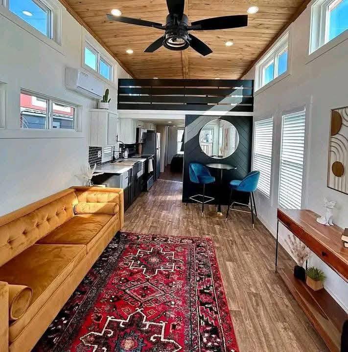 Modern tiny home interior with a cozy yellow couch, vibrant red rug, and stylish kitchen. Features a loft, wooden ceiling, and blue dining chairs.