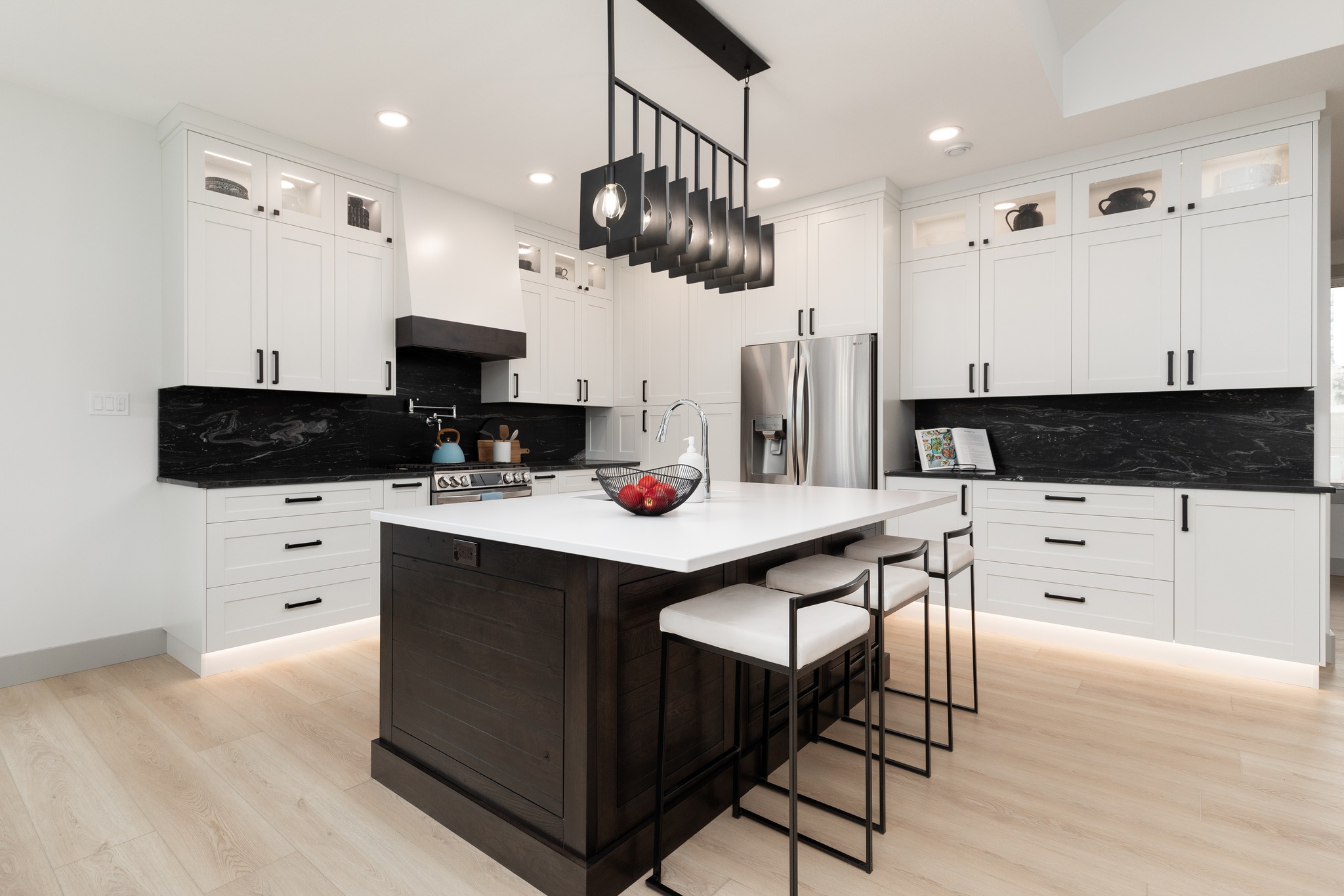 Sleek modern kitchen with white cabinets, black marble backsplash, large island with dark wood base, minimalist pendant lights, and light wood flooring.
