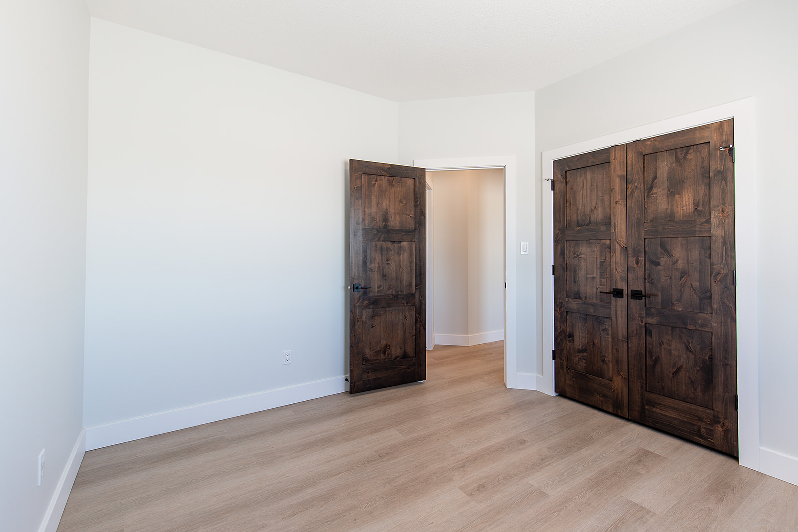 A minimalist empty room with light wood flooring and white walls, featuring an open wooden door on the left and closed double closet doors on the right.