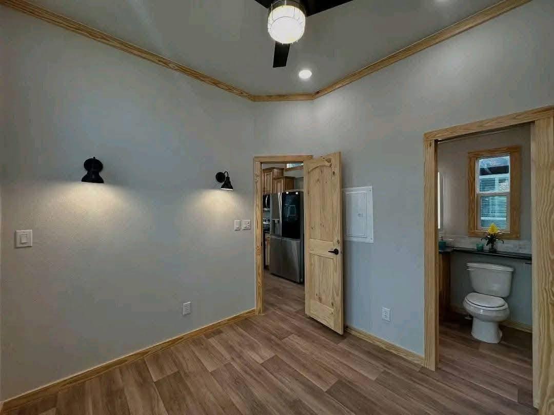 A small, empty room with wooden trim and laminate flooring features wall sconces, an open door revealing stainless steel appliances, and a view into a bathroom.