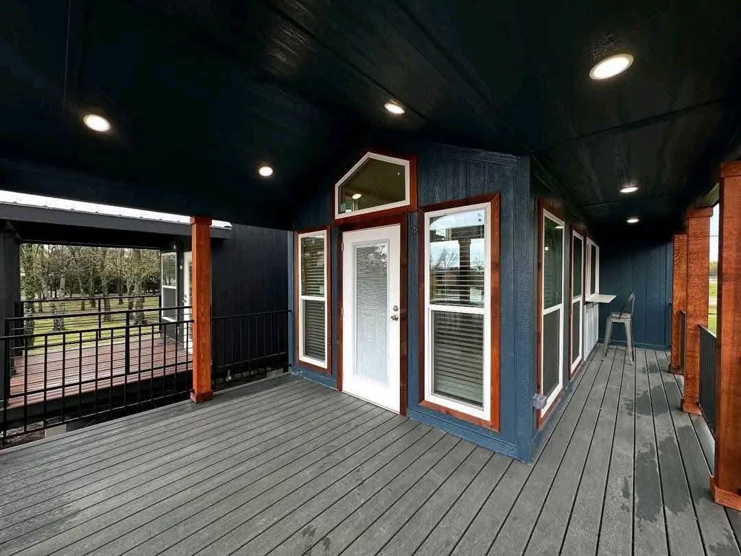 Covered porch with dark wood siding and a white door surrounded by windows. Recessed lighting creates a cozy atmosphere. Trees visible outside.