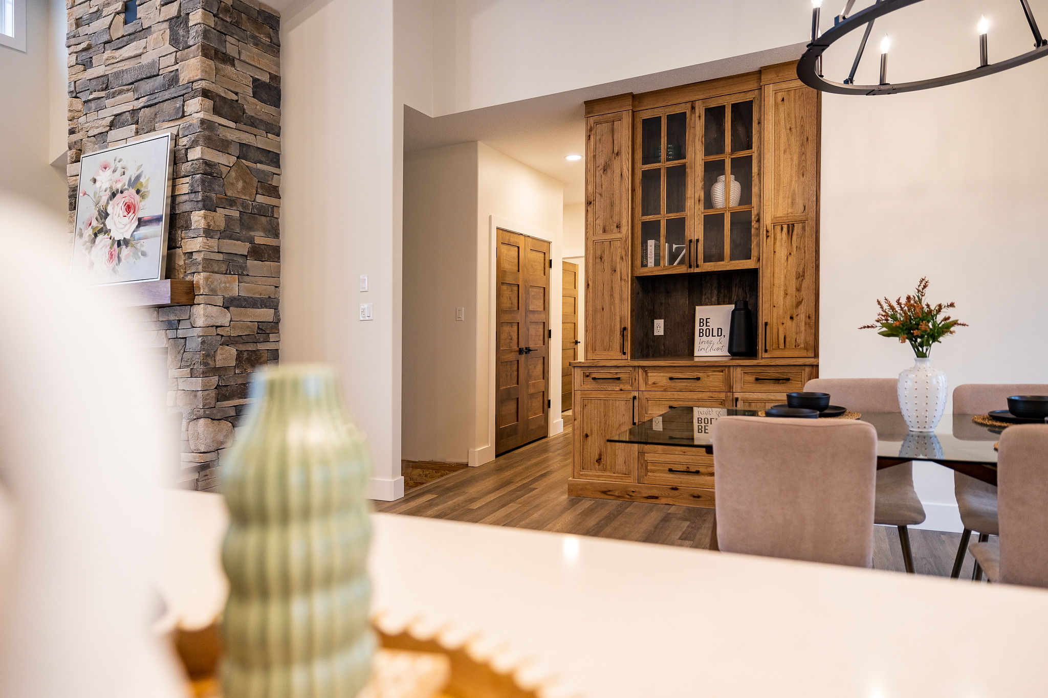 Cozy living room with a stone fireplace, floral art, and wooden cabinet. A glass dining table with plush chairs and a modern chandelier enhance warmth.