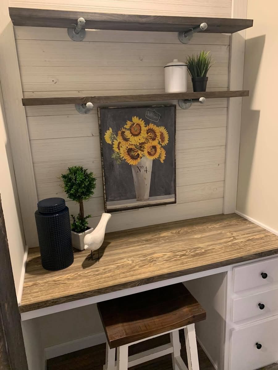 Cozy nook with wood desk, two shelves above. Features a sunflower painting, plants, a white jar, blue vase, and white bird figurine. Rustic charm.