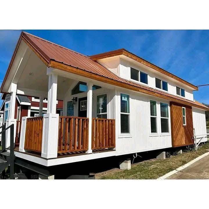 A modern tiny house with a wooden porch and white exterior. It features a slanted red roof and multiple windows, set against a clear blue sky.