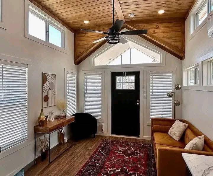 Cozy living room with a wooden vaulted ceiling, black ceiling fan, large black door, and natural light from windows. Features a sofa, rug, and art.