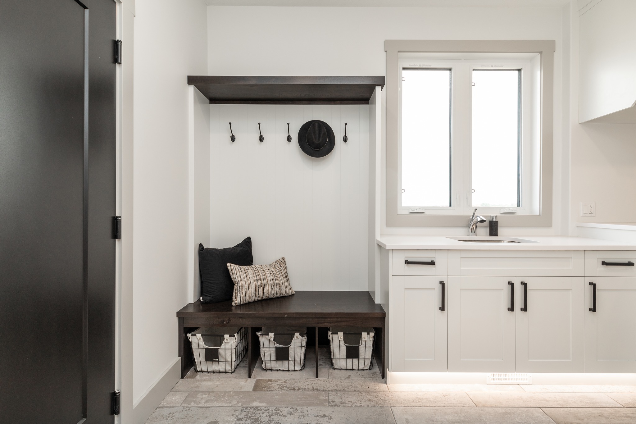 A modern mudroom with white walls, featuring a dark wooden bench with cushions, hooks above holding a hat, and three storage baskets below. Bright, minimalistic ambiance.