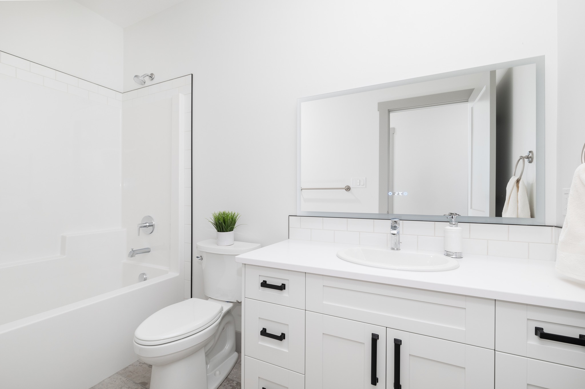 A modern, white bathroom with a sleek sink and faucet, large mirror, white cabinets with black handles, and a white bathtub. A small plant adds greenery.