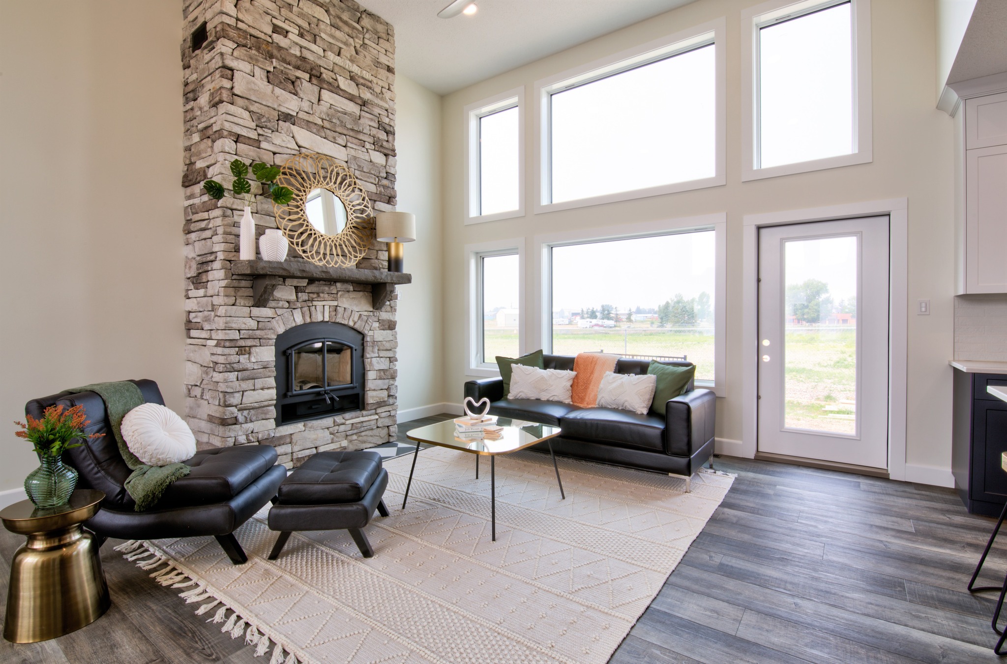 Modern living room with a tall stone fireplace, black leather seating, large windows, and a glass coffee table on a beige rug, creating a cozy ambiance.