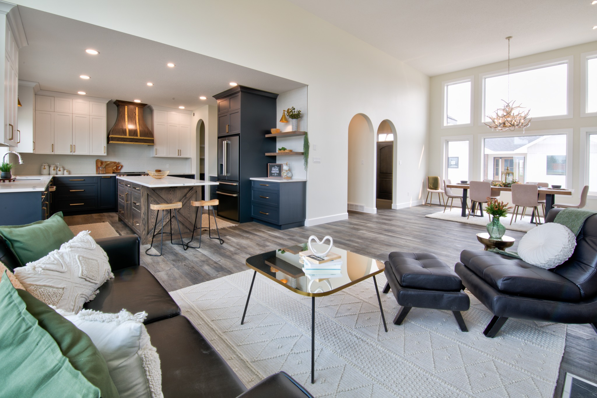 Spacious modern living room and kitchen with neutral tones. Features include black and white cabinetry, wood flooring, a cozy seating area, and a chandelier-lit dining space.