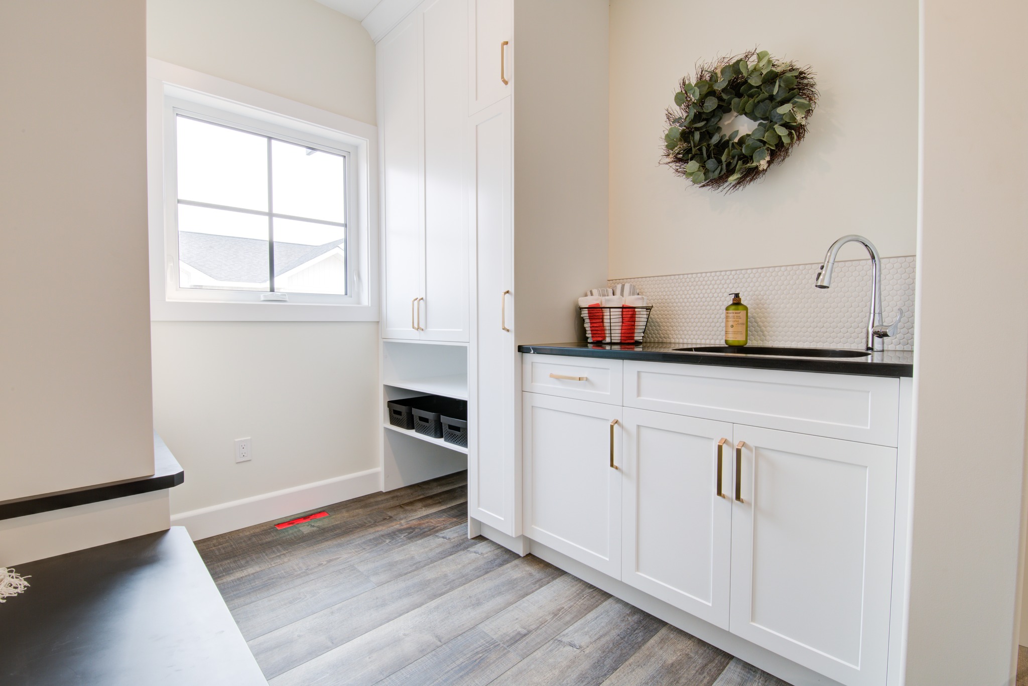 Bright laundry room with white cabinetry, dark countertop, and wood-look flooring. A window lets in natural light. A green wreath adds a touch of decor.