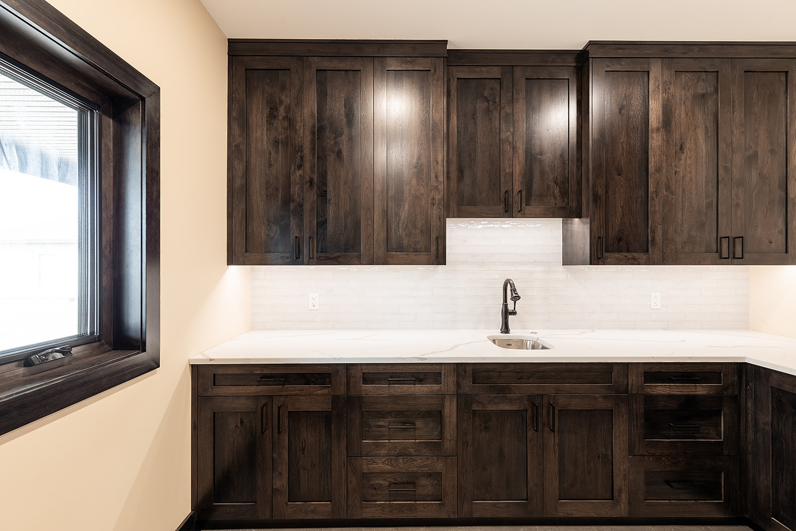 A modern kitchen with dark wooden cabinets, white countertops, and a small sink. The room has a warm, inviting feel with ample natural light.