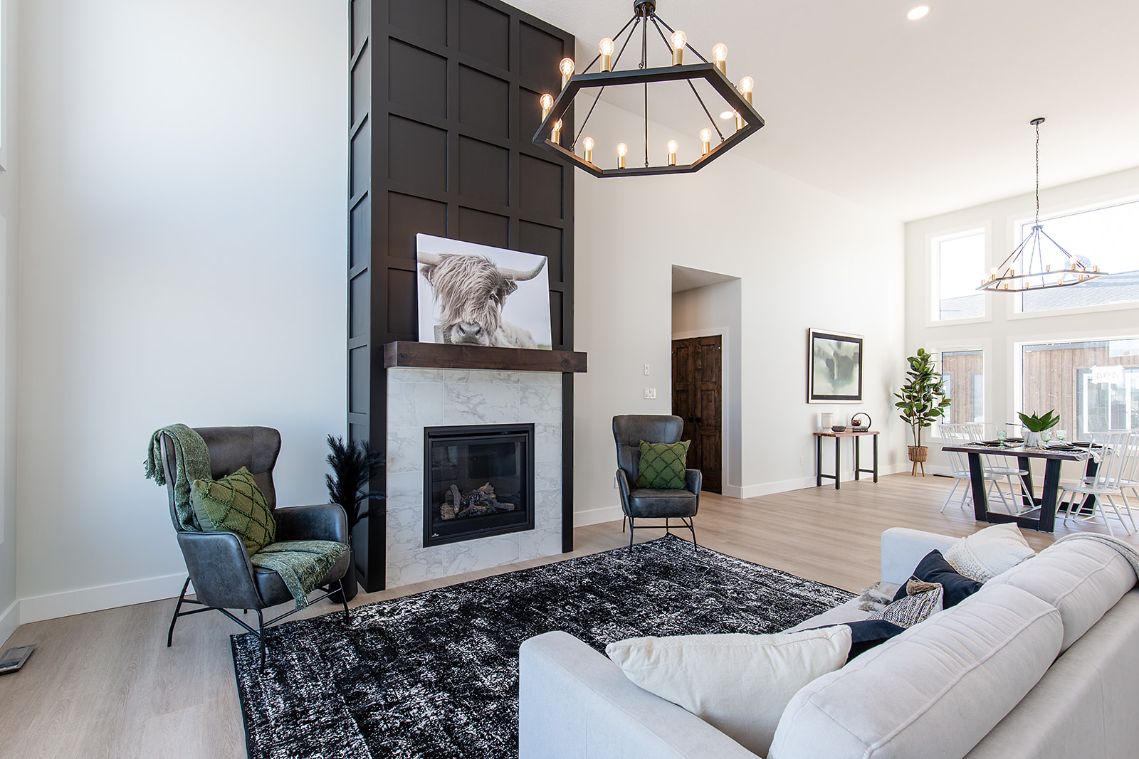 Modern living room with high ceiling, featuring a marble fireplace, dark geometric mantel, and a cow portrait. The area has plush seating, a black rug, and large windows.