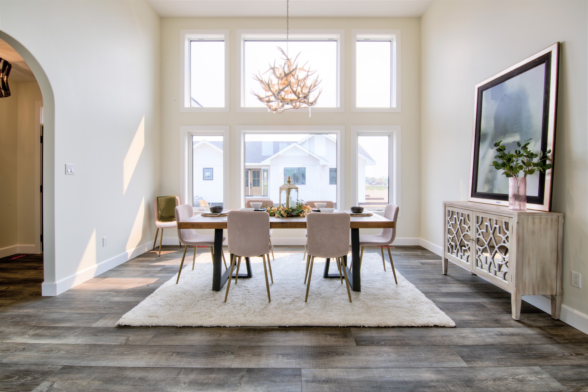 Bright dining room with a large window, modern chandelier, and a wooden table set for four. Decor includes a sideboard with foliage. Cozy and elegant ambiance.