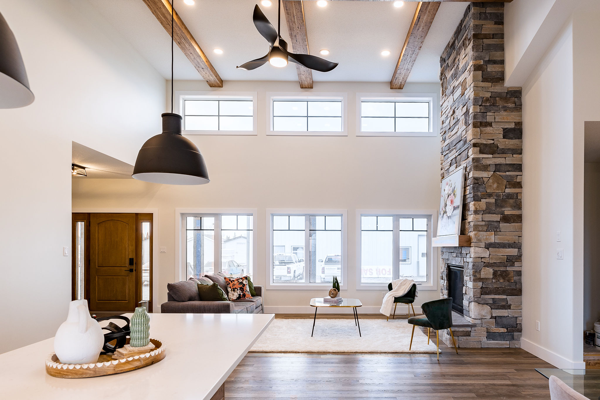 Spacious living room with high ceiling, exposed wood beams, and large windows. Stone fireplace, cozy seating, modern decor, and soft lighting.
