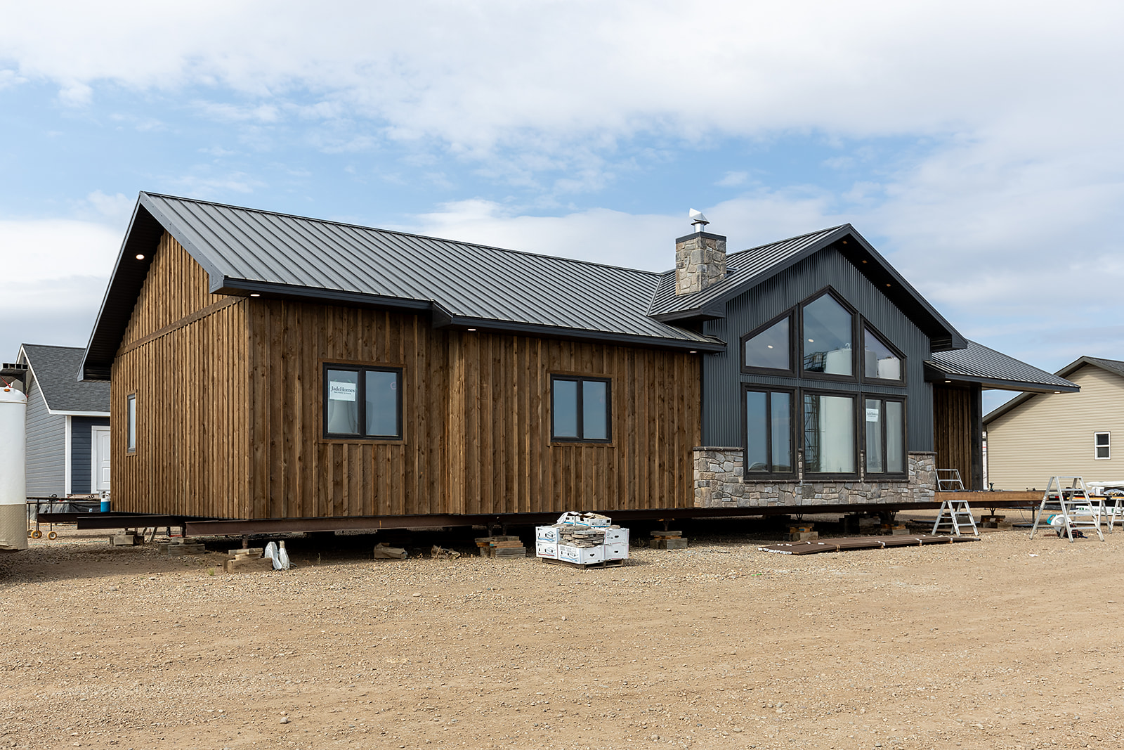A modern wooden house with large windows, set on blocks above a dirt lot under a partly cloudy sky, suggesting construction or relocation.