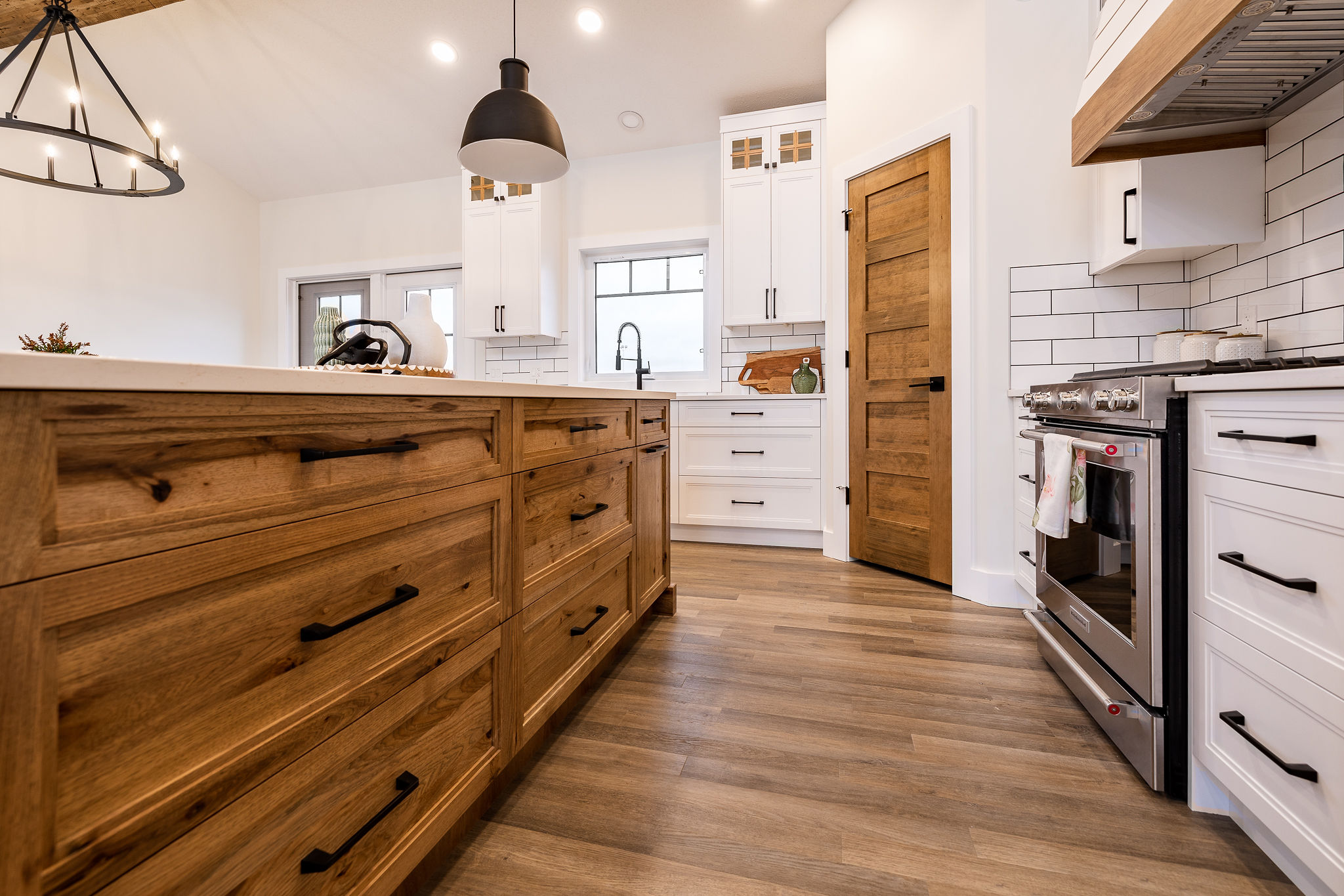 Modern kitchen with wooden cabinets, white tiled walls, and hardwood floors. Features a large island, stainless steel stove, and pendant lighting. Warm, inviting tone.