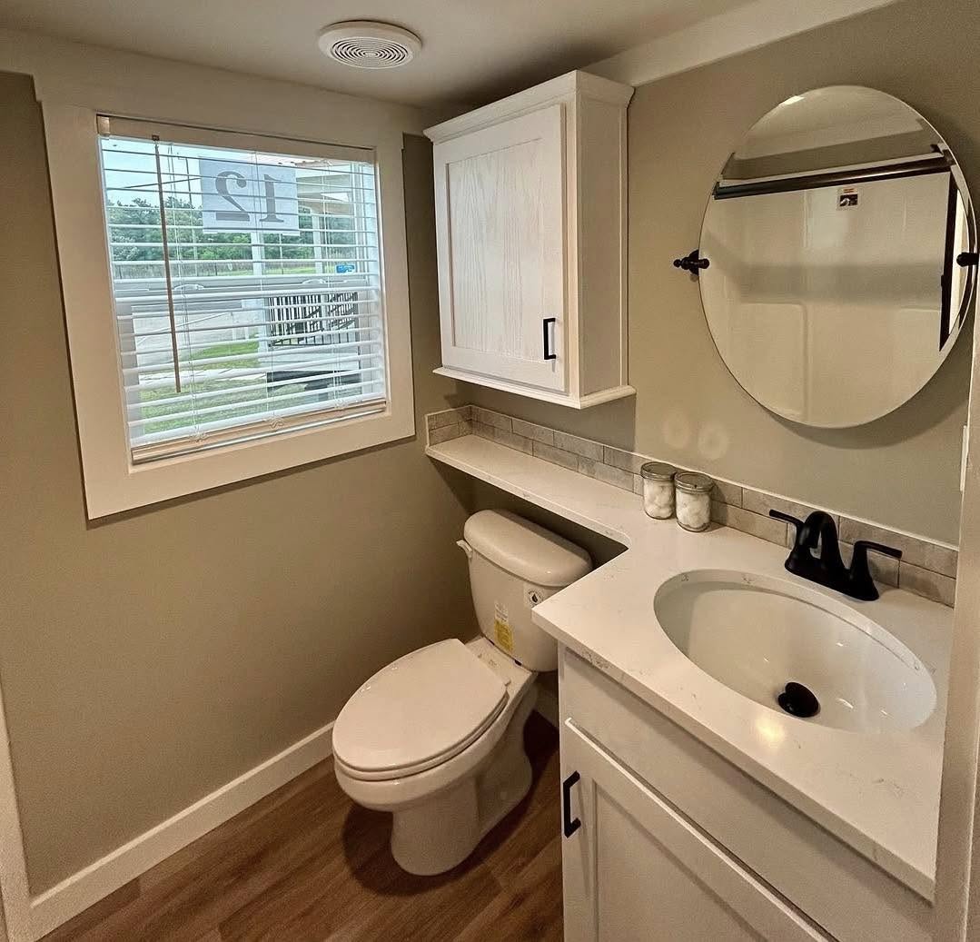 A small bathroom with light brown walls, featuring a window with blinds, white cabinets, a round mirror, and a white sink and toilet.