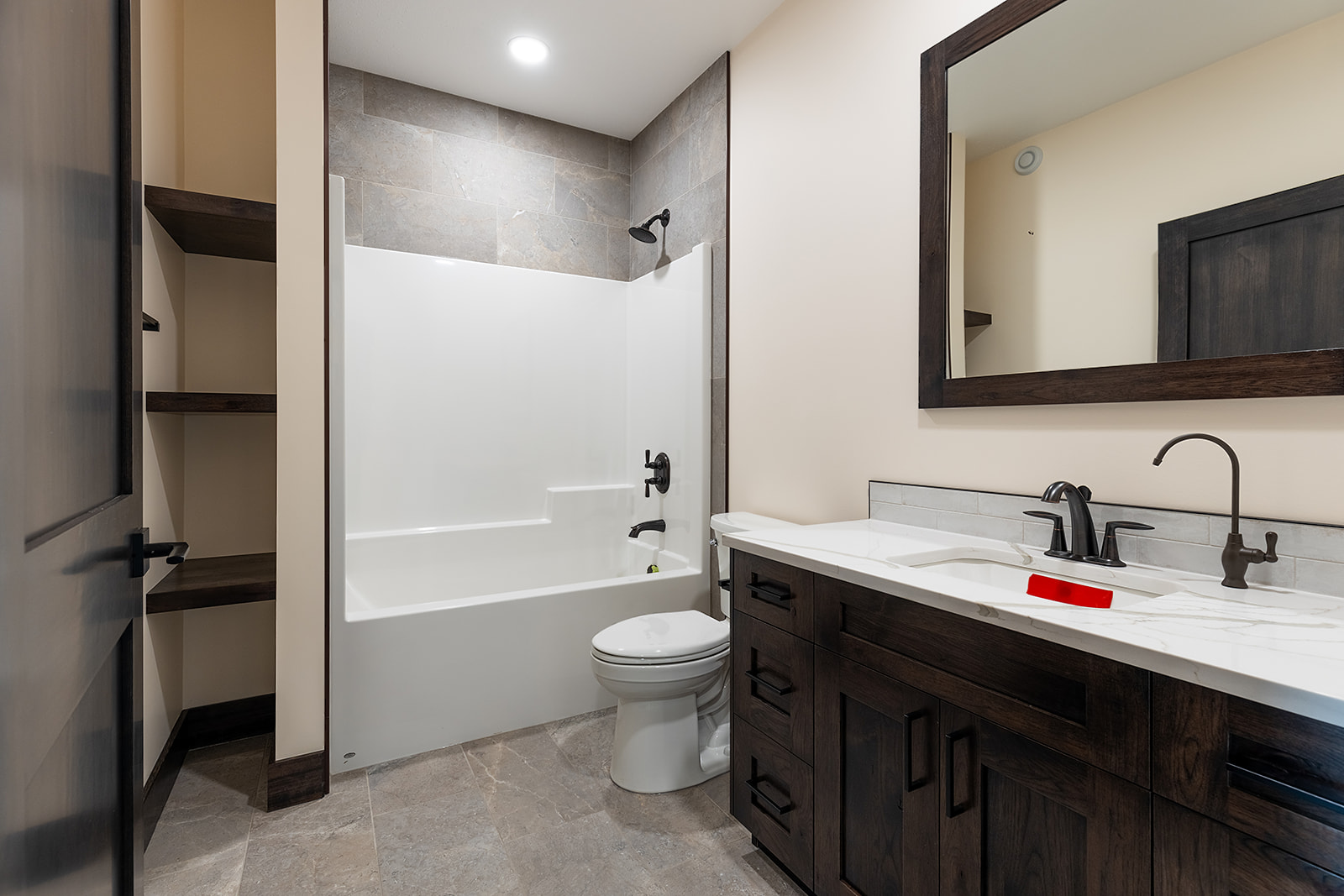Modern bathroom features a white bathtub with a dark showerhead, a toilet, wooden shelves, and a dark wood vanity with a large mirror, conveying a clean, minimalist design.