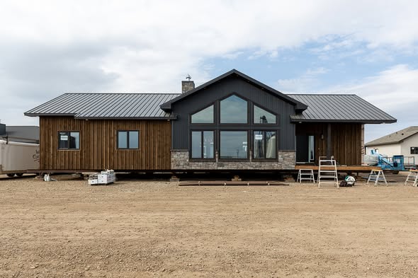 A modern, single-story house with a gable roof and large central windows, surrounded by a barren dirt lot under a cloudy sky. Construction tools are visible.