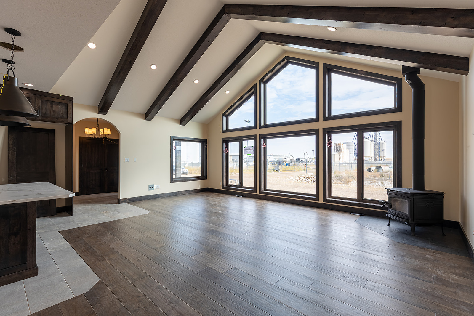 Spacious living room with vaulted ceiling and exposed beams, large geometric windows offering natural light, hardwood floors, and a wood stove.