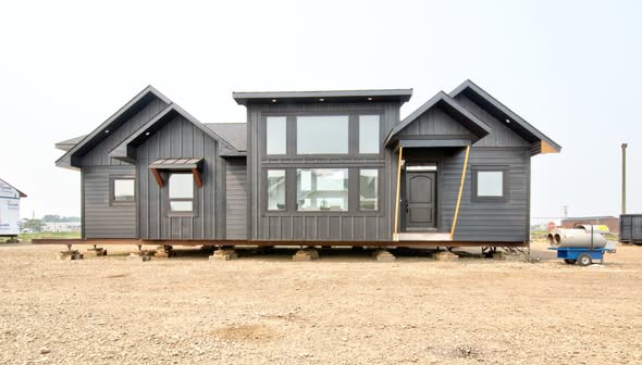 Dark gray modular home with A-frame peaks, large windows, and a central door. The home is on stilts in a barren, dirt-covered landscape.