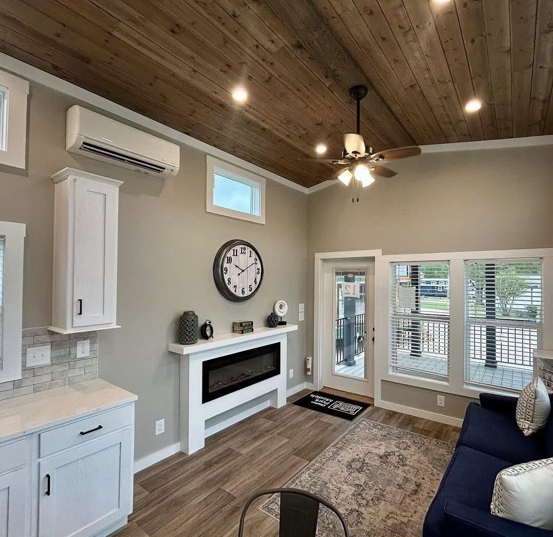 Stylish living room with wood ceiling, fan, and modern decor. Features a clock above a sleek fireplace, large windows, and cozy seating. Warm ambiance.