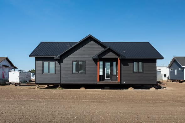 Black modular home with a pitched roof sits on a dry, dirt lot under a clear blue sky. The house has large windows and a front door with glass panels.