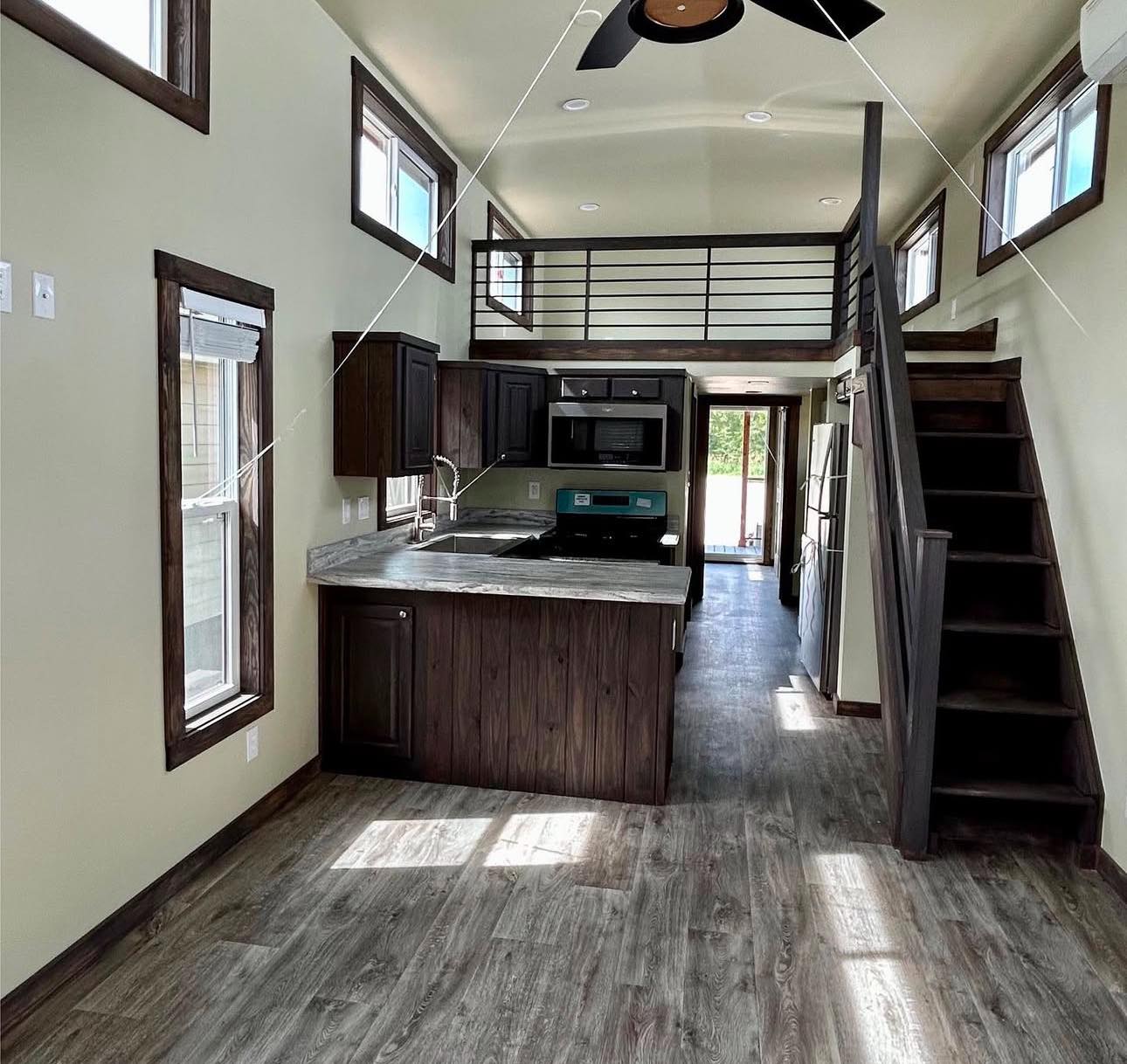 A small, modern tiny house kitchen features dark wood cabinetry, a loft area with railing, light walls, wooden stairs, and a ceiling fan.