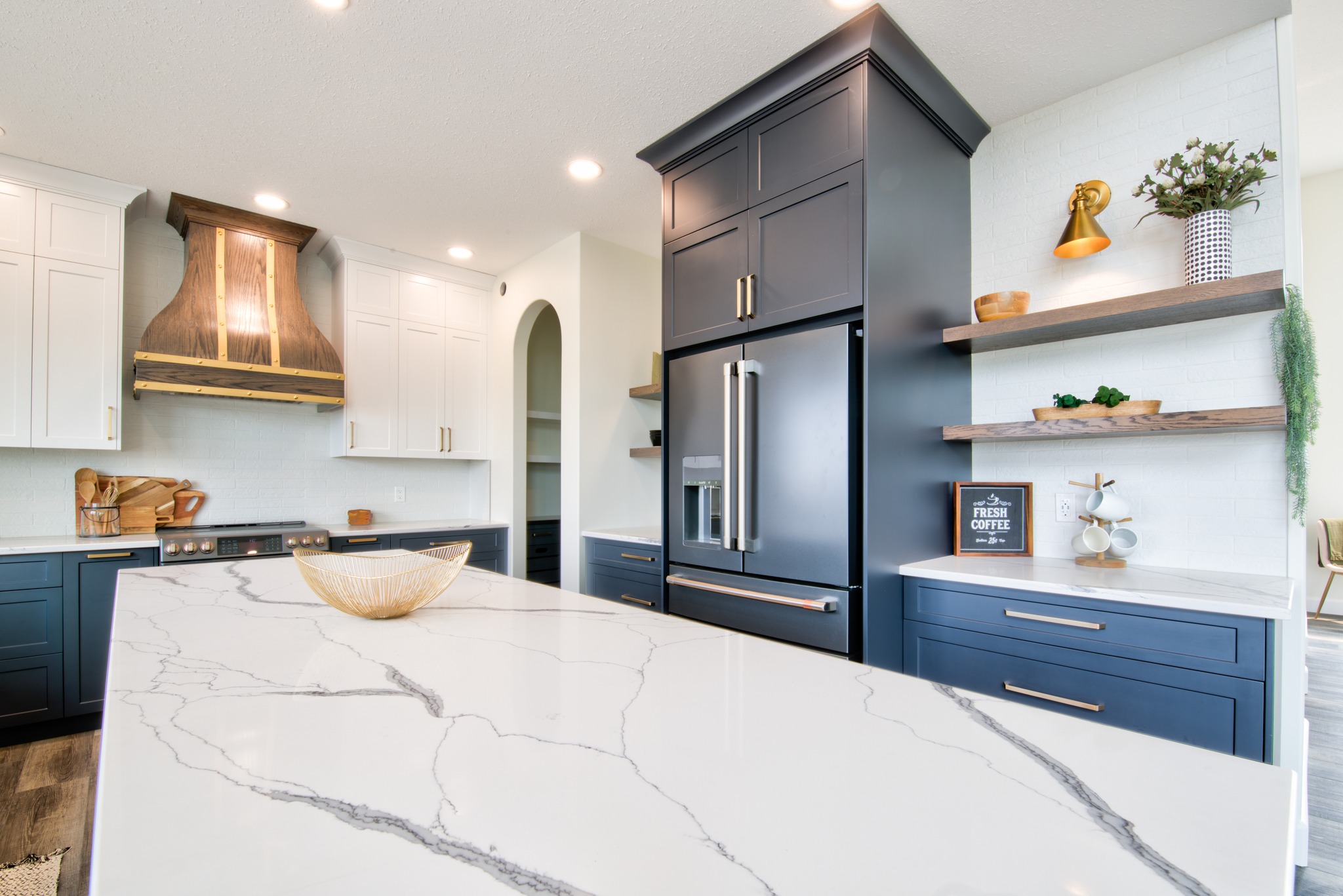 Modern kitchen with blue and white cabinetry, a large marble island, bronze accents, and open shelves. A cozy and elegant ambiance.